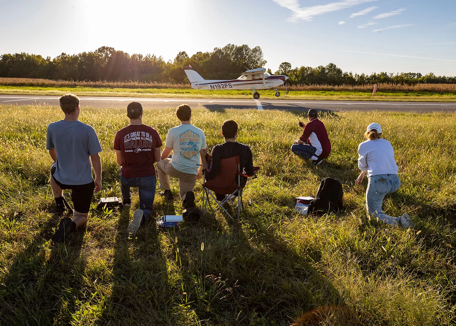Members of the Flying Salukis sit in a grassy area near an airport runway and watch a small, SIU-branded plane land.